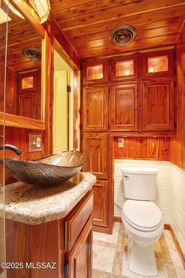 half bathroom featuring vanity, visible vents, toilet, wooden ceiling, and wooden walls