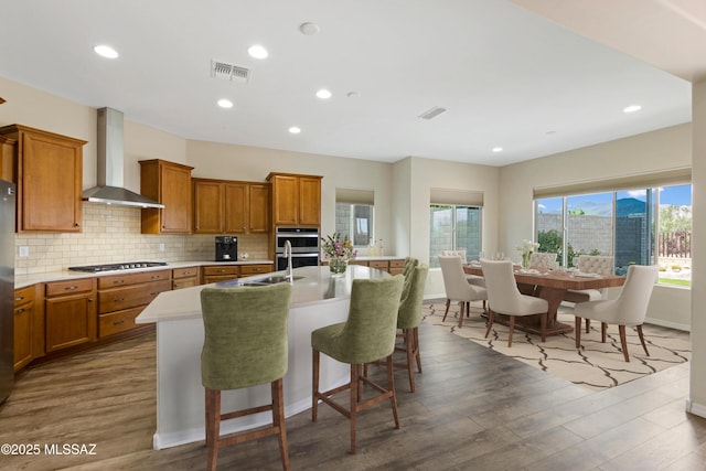 13906 N Rim Trl, Marana, AZ 85658 - kitchen with wall chimney exhaust hood, stainless steel gas stovetop, brown cabinets, an island with sink, and dark wood finished floors