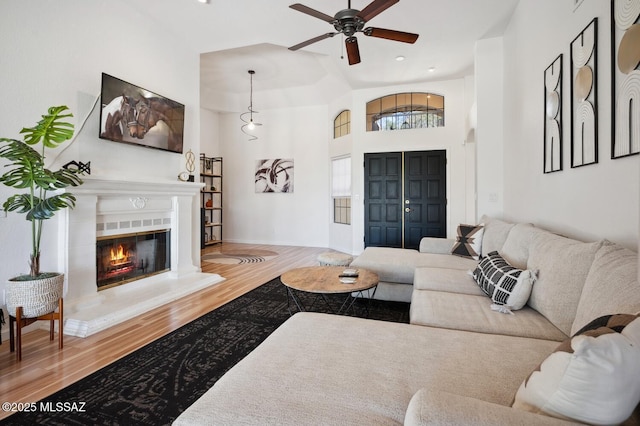 living area featuring a glass covered fireplace, a towering ceiling, ceiling fan, and wood finished floors
