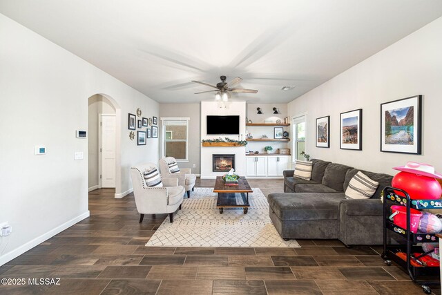 living area featuring wood finish floors, arched walkways, a ceiling fan, a glass covered fireplace, and baseboards