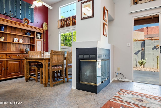 dining area featuring a multi sided fireplace, a ceiling fan, tile patterned flooring, a high ceiling, and baseboards