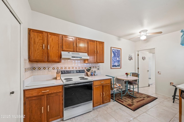 kitchen featuring tasteful backsplash, light countertops, under cabinet range hood, brown cabinetry, and white electric range oven