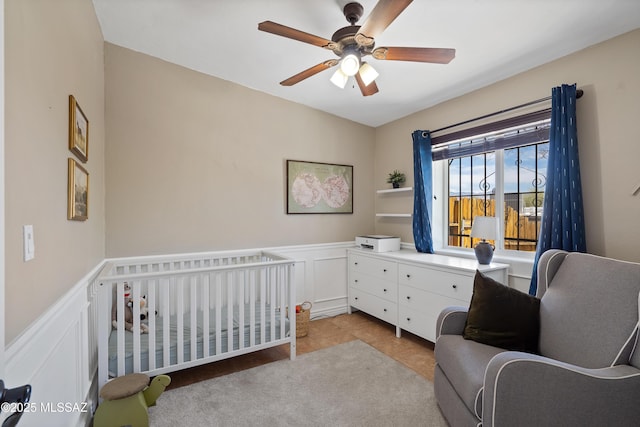 bedroom with a nursery area, wainscoting, a decorative wall, ceiling fan, and tile patterned flooring