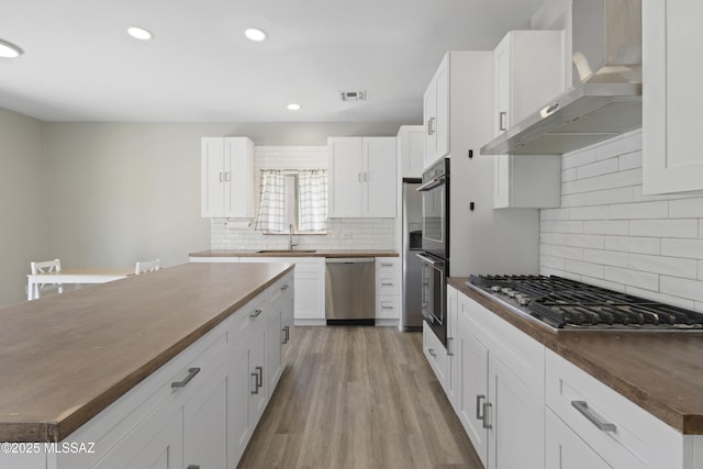 kitchen featuring wall chimney range hood, tasteful backsplash, a sink, wood counters, and appliances with stainless steel finishes