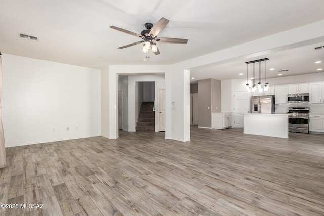 unfurnished living room featuring stairs, visible vents, light wood-style floors, and ceiling fan