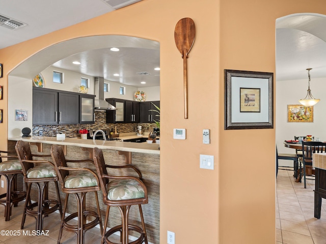 kitchen featuring arched walkways, a breakfast bar, wall chimney range hood, visible vents, and light tile patterned flooring