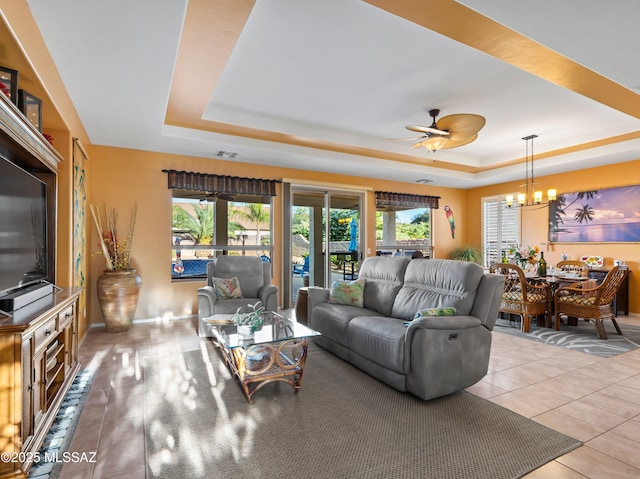 tiled living room with ceiling fan with notable chandelier, visible vents, a raised ceiling, and a healthy amount of sunlight