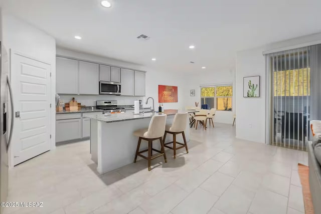 kitchen with stainless steel appliances, a sink, a center island with sink, a kitchen bar, and gray cabinetry