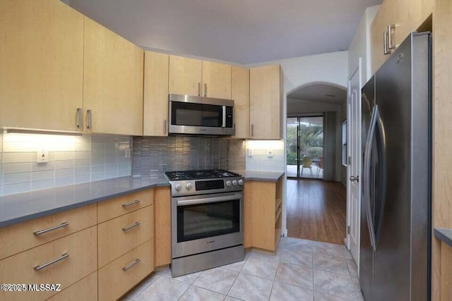 kitchen featuring light brown cabinetry, stainless steel appliances, light tile patterned floors, arched walkways, and tasteful backsplash