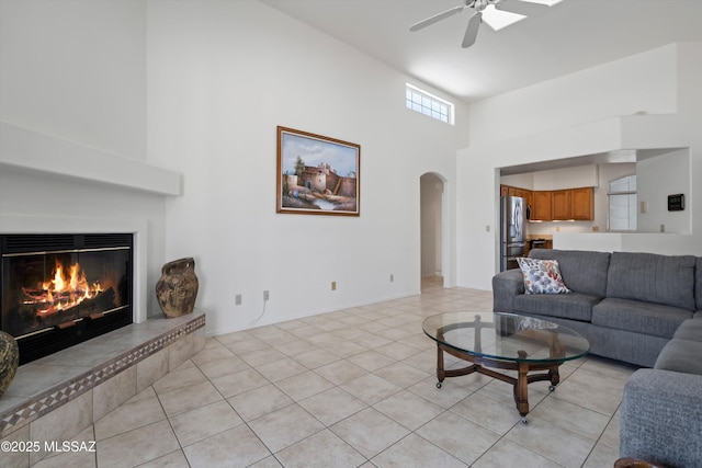 living area with arched walkways, light tile patterned floors, a glass covered fireplace, a ceiling fan, and a towering ceiling