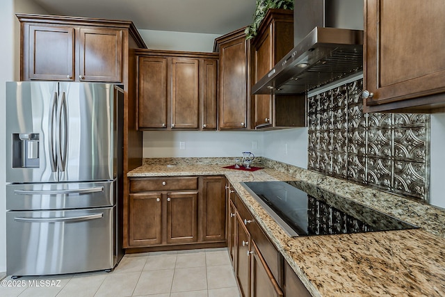 kitchen featuring light stone countertops, light tile patterned flooring, black electric stovetop, stainless steel fridge, and wall chimney exhaust hood