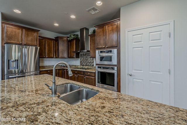 kitchen with light stone countertops, recessed lighting, wall chimney exhaust hood, a sink, and stainless steel appliances