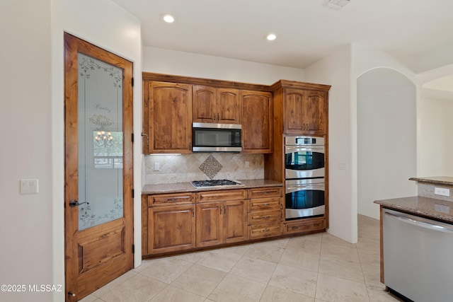 kitchen featuring dark stone counters, stainless steel appliances, brown cabinets, and decorative backsplash