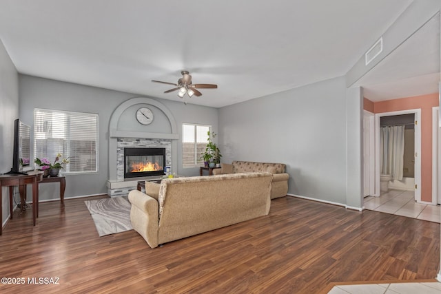 living room with wood finished floors, ceiling fan, a stone fireplace, baseboards, and visible vents