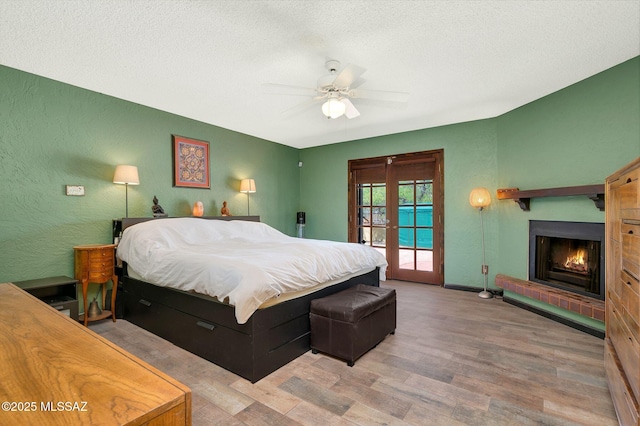 bedroom with a textured ceiling, a warm lit fireplace, access to exterior, wood finished floors, and a ceiling fan