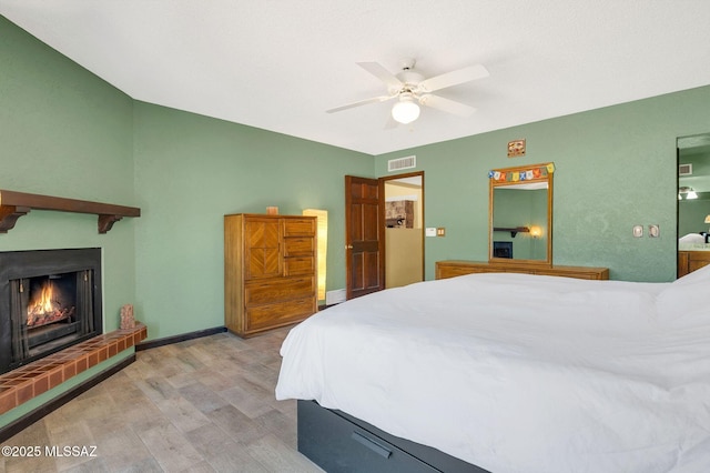 bedroom featuring a warm lit fireplace, light wood-style flooring, ceiling fan, baseboards, and visible vents