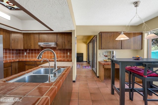 kitchen featuring under cabinet range hood, brown cabinets, backsplash, a sink, and a textured ceiling