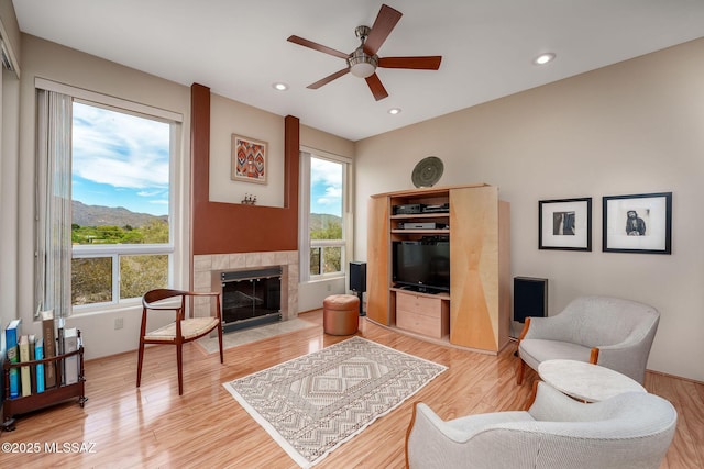 living area with a wealth of natural light, a ceiling fan, a tiled fireplace, and light wood finished floors