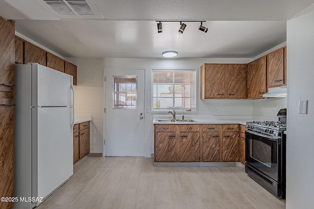 kitchen with a sink, black gas range oven, freestanding refrigerator, under cabinet range hood, and brown cabinetry