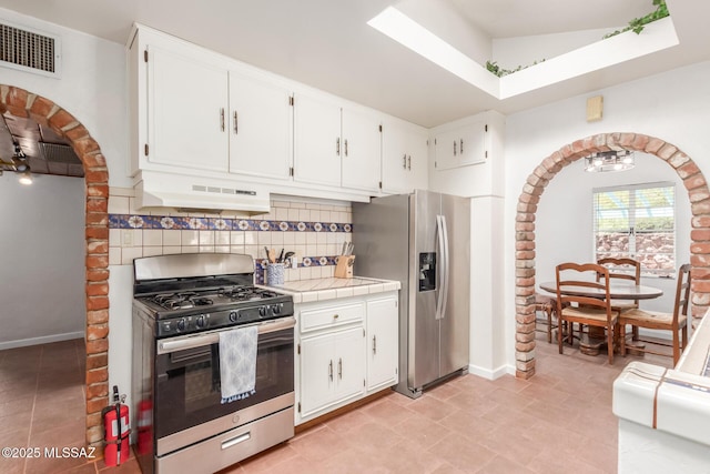 kitchen with stainless steel appliances, under cabinet range hood, backsplash, tile countertops, and white cabinetry