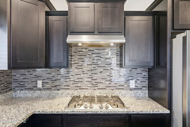 kitchen with dark brown cabinets, decorative backsplash, appliances with stainless steel finishes, under cabinet range hood, and light stone countertops