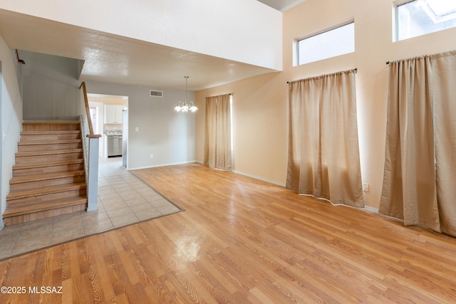 unfurnished living room featuring light wood-type flooring, a towering ceiling, visible vents, a notable chandelier, and stairway