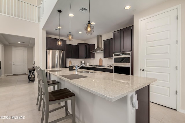 kitchen featuring wall chimney range hood, light stone counters, a kitchen breakfast bar, a sink, and appliances with stainless steel finishes