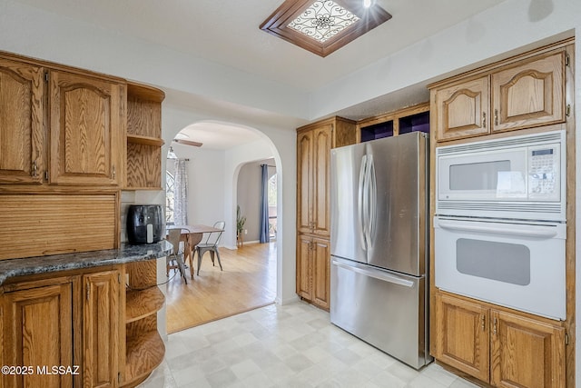 kitchen with open shelves, brown cabinetry, white appliances, dark countertops, and arched walkways