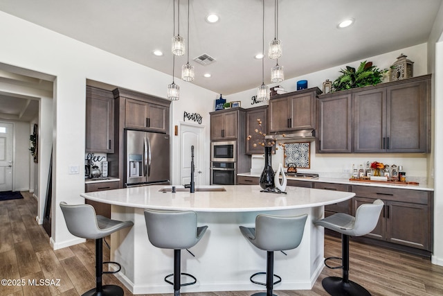 kitchen featuring an island with sink, dark brown cabinetry, stainless steel appliances, dark wood finished floors, and visible vents