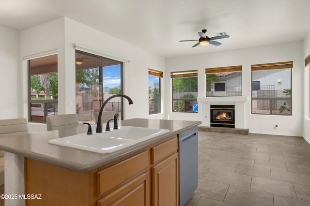 kitchen featuring a kitchen island with sink, a sink, ceiling fan, a glass covered fireplace, and dishwasher