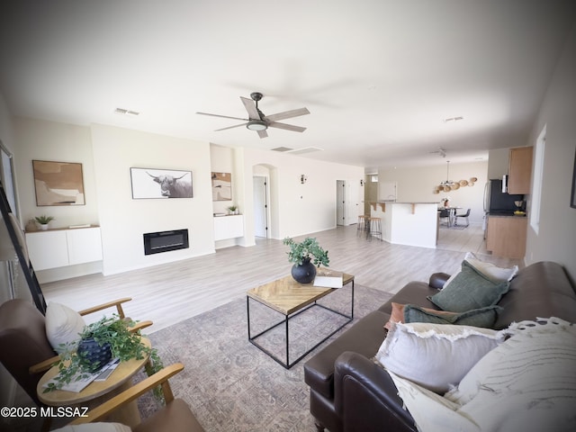 living area with visible vents, a glass covered fireplace, light wood-style floors, ceiling fan, and arched walkways