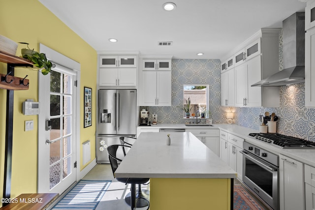 kitchen with wall chimney range hood, appliances with stainless steel finishes, a sink, backsplash, and a kitchen island