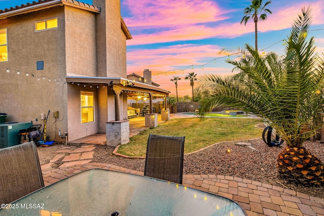 back of house at dusk featuring fence, stucco siding, a chimney, a patio, and a yard