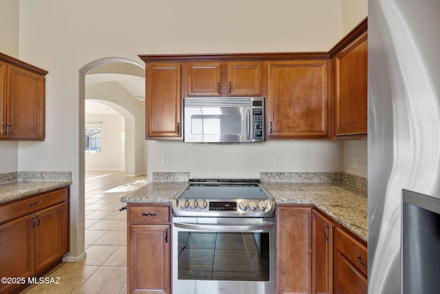 kitchen featuring arched walkways, light stone countertops, brown cabinets, light tile patterned floors, and stainless steel appliances