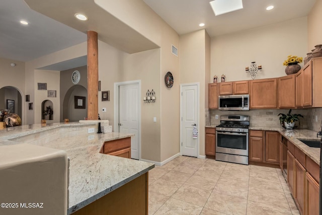 kitchen featuring brown cabinetry, a high ceiling, tasteful backsplash, arched walkways, and appliances with stainless steel finishes