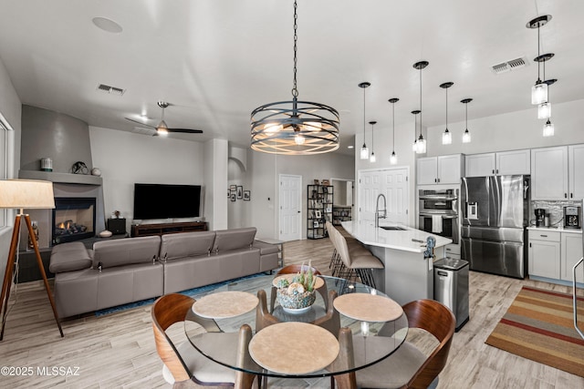 dining space featuring light wood finished floors, visible vents, a large fireplace, and ceiling fan with notable chandelier