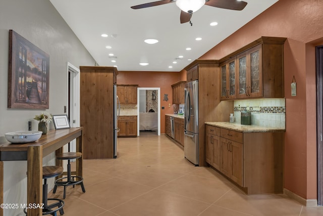 kitchen featuring brown cabinetry, freestanding refrigerator, glass insert cabinets, a ceiling fan, and backsplash