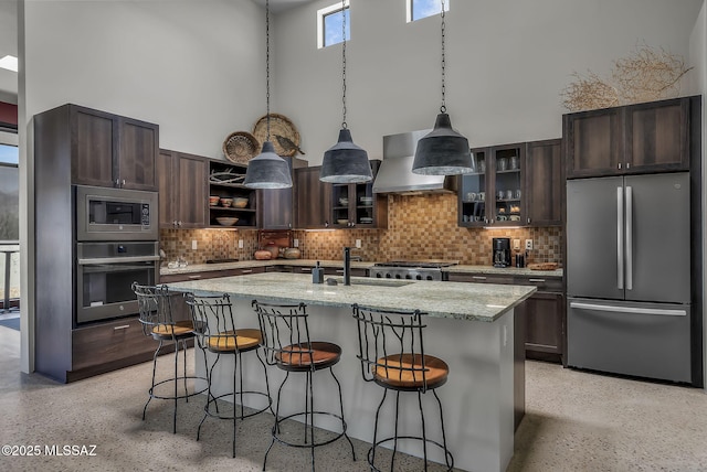 kitchen with a high ceiling, appliances with stainless steel finishes, a sink, wall chimney exhaust hood, and light speckled floor