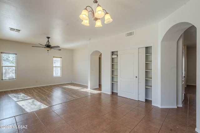 tiled empty room featuring arched walkways, ceiling fan with notable chandelier, built in shelves, and visible vents