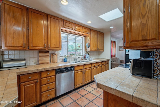 kitchen featuring brown cabinets, a sink, tasteful backsplash, stainless steel dishwasher, and a skylight