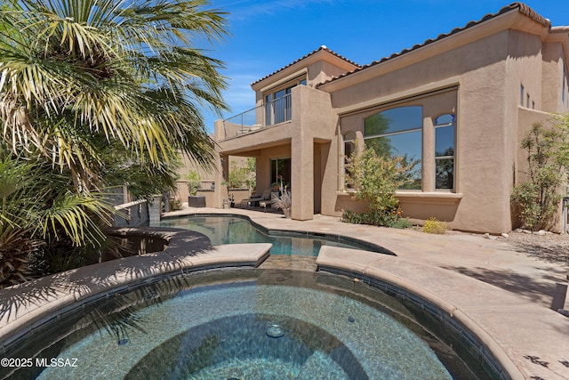 rear view of property featuring an in ground hot tub, an outdoor pool, a patio area, a balcony, and stucco siding