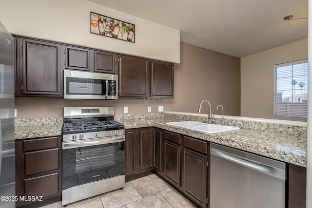 kitchen featuring dark brown cabinetry, light tile patterned flooring, light stone countertops, a sink, and appliances with stainless steel finishes