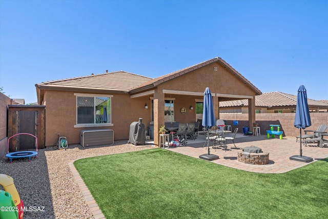 back of house featuring a trampoline, stucco siding, a patio, a yard, and a fire pit