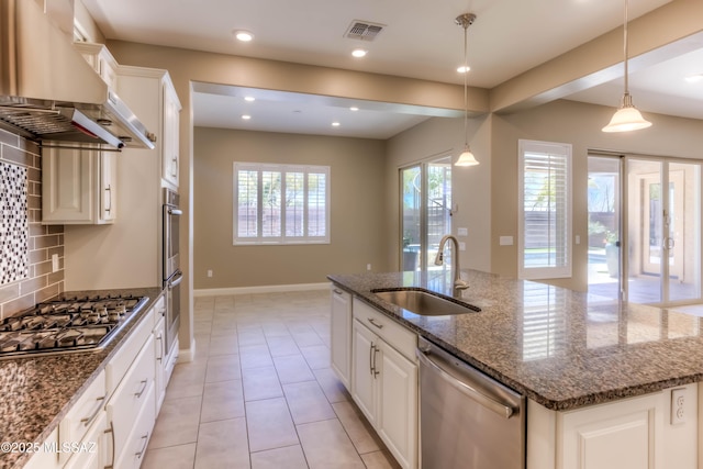 kitchen with a sink, wall chimney exhaust hood, stainless steel appliances, a center island with sink, and visible vents