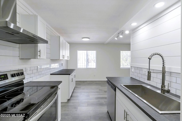 kitchen with dark countertops, appliances with stainless steel finishes, a sink, wall chimney exhaust hood, and backsplash