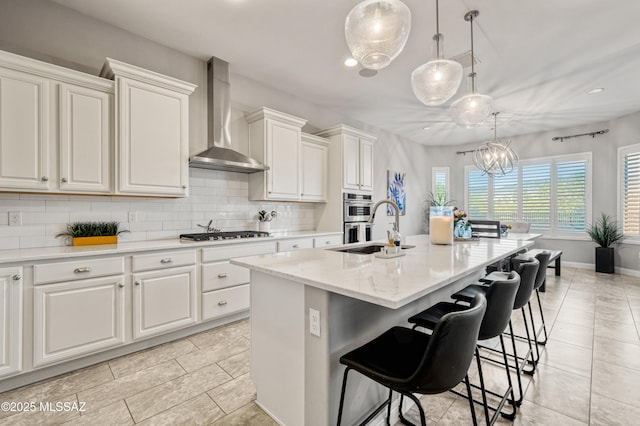 kitchen with a kitchen island with sink, wall chimney range hood, gas stovetop, white cabinets, and tasteful backsplash