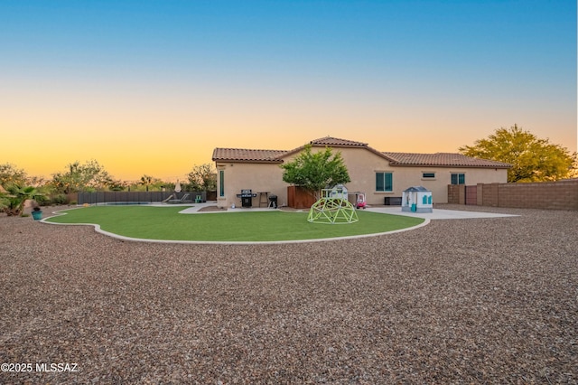 back of property at dusk featuring a tile roof, a lawn, a patio area, a fenced backyard, and stucco siding
