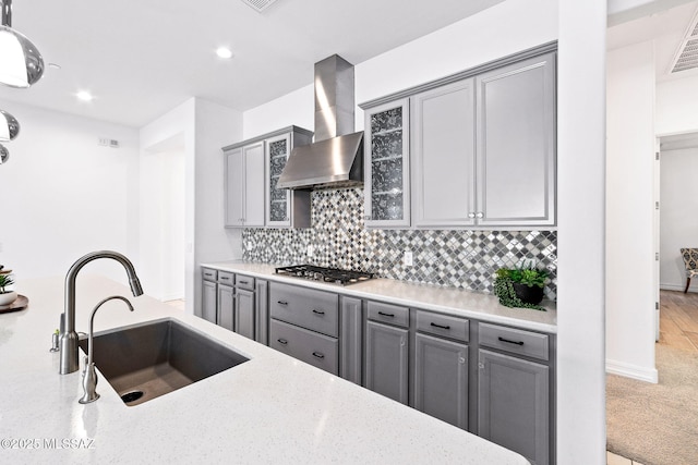 kitchen with glass insert cabinets, wall chimney range hood, gray cabinets, tasteful backsplash, and a sink