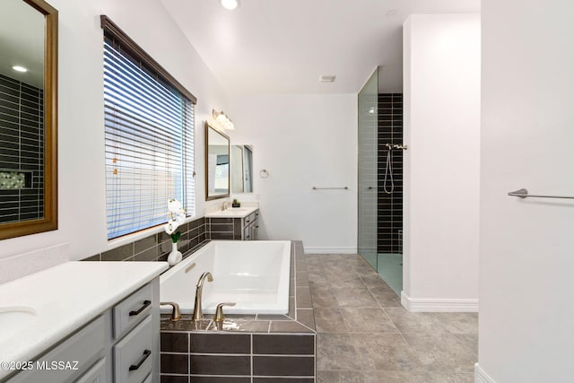 bathroom with a garden tub, baseboards, tiled shower, two vanities, and a sink