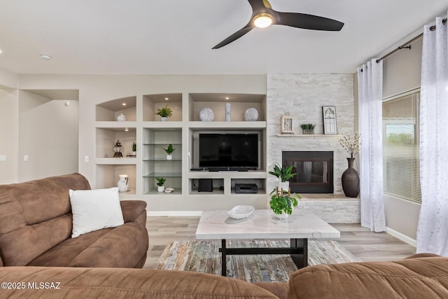 living area with baseboards, a ceiling fan, light wood finished floors, and a stone fireplace